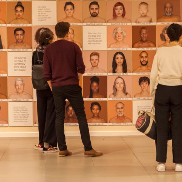 The image shows a group of people observing an exhibition at the Migration Museum. The exhibition shows numerous faces on a wall.