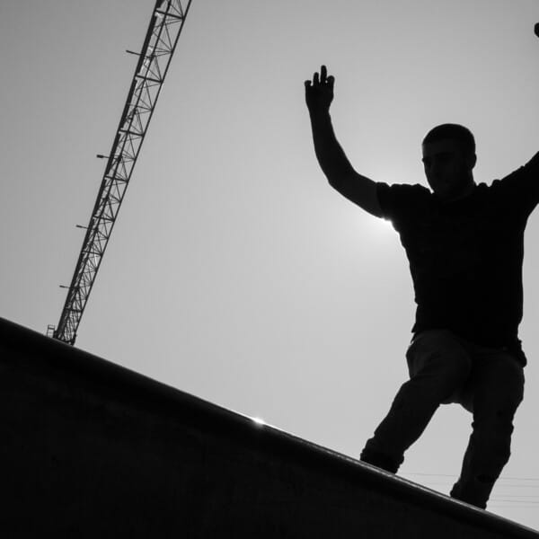 A black and white image, showing the silhouette of a person standing on a roof with their hands raised above their head.
