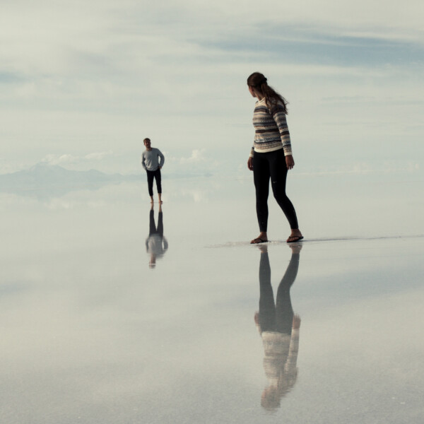 Two human beings walk across a shallow span of water and sand. They are reflected clearly in the water.