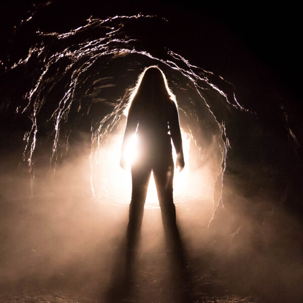 A person stands in a dark cave, looking towards light shining from the opening.