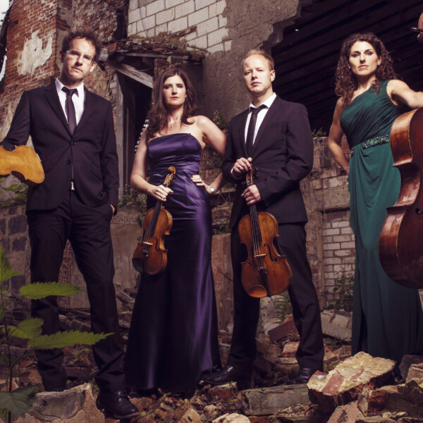The Carducci Quartet stand upon brick rubble in concert dress and holding their stringed instruments in hand.
