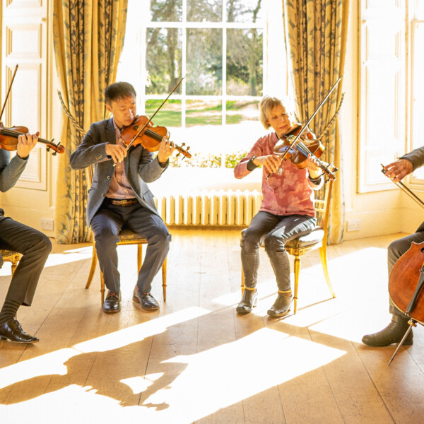 The four members of the Edinburgh Quartet are sat in a sunlit room, playing their respective instruments.
