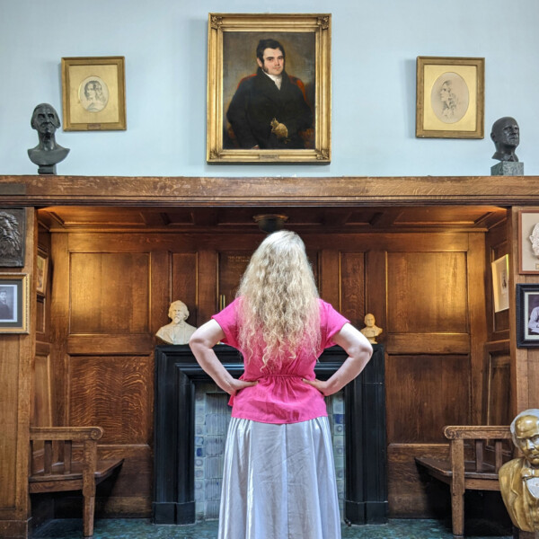 Frances M Lynch, Artistic Director of Electric Voice Theatre, stands in Conway Hall's library. With her back to the camera, she looks up towards two portraits of the Flower sisters.